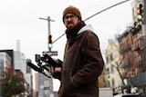John Wilson stands looking concerned and rugged up on a NYC street, camera in hand.