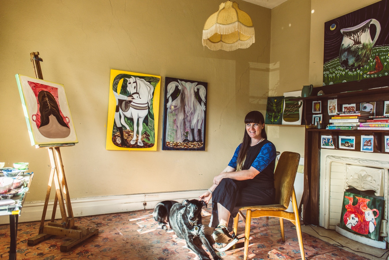 A woman and her dog sit in studio full of paintings and books