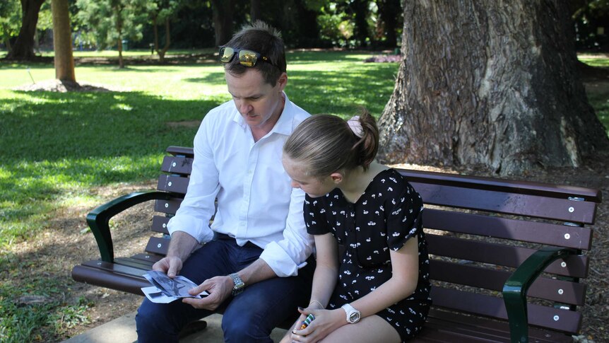 A father and daughter sit on a park bench looking at war medals.