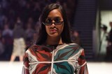 A woman wearing sunglasses poses on a fashion runway.
