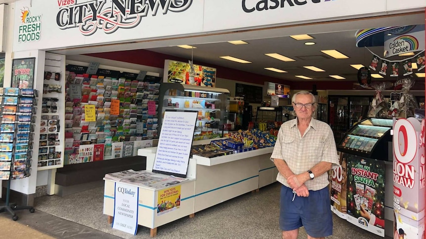 A man with grey hair stands outside a newsagency. He is wearing a grey cheque shirt and navy shorts.