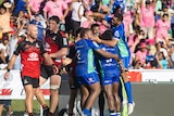 Rugby players wearing blue, huddle in celebration after scoring, while two opposition players in red look exhausted