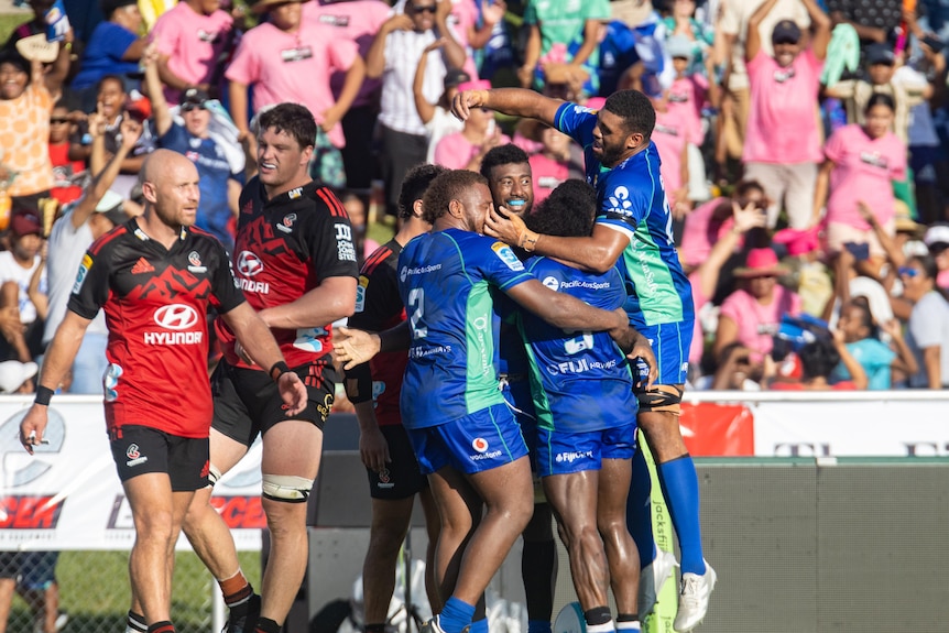 Rugby players wearing blue, huddle in celebration after scoring, while two opposition players in red look exhausted