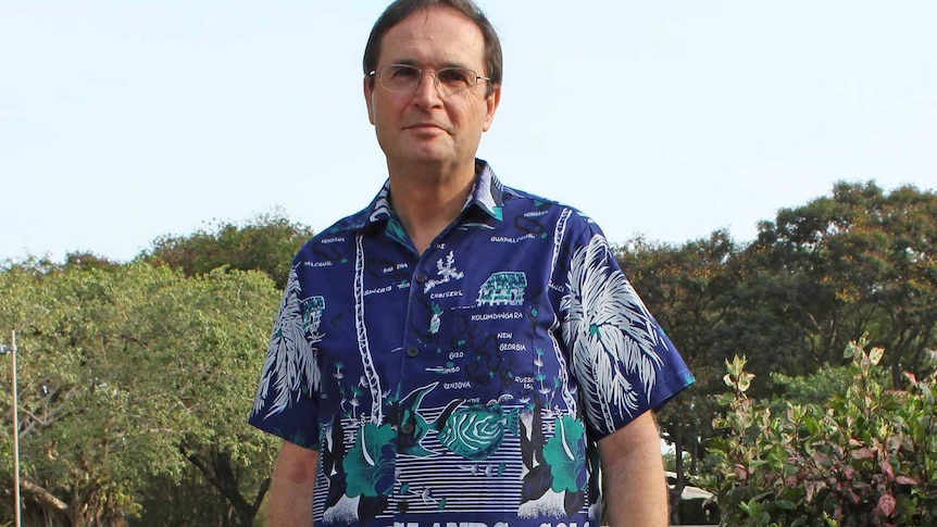 A middle-aged male reverend stands in sandals and a print t-shirt with the words Solomon Islands written across it.