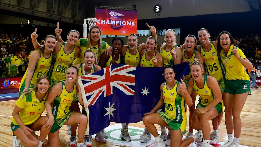 A netball team gathers on court for a group shot holding an Australian flag as a sign says 'Champions' behind them.