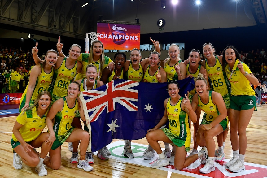 A netball team gathers on court for a group shot holding an Australian flag as a sign says 'Champions' behind them.