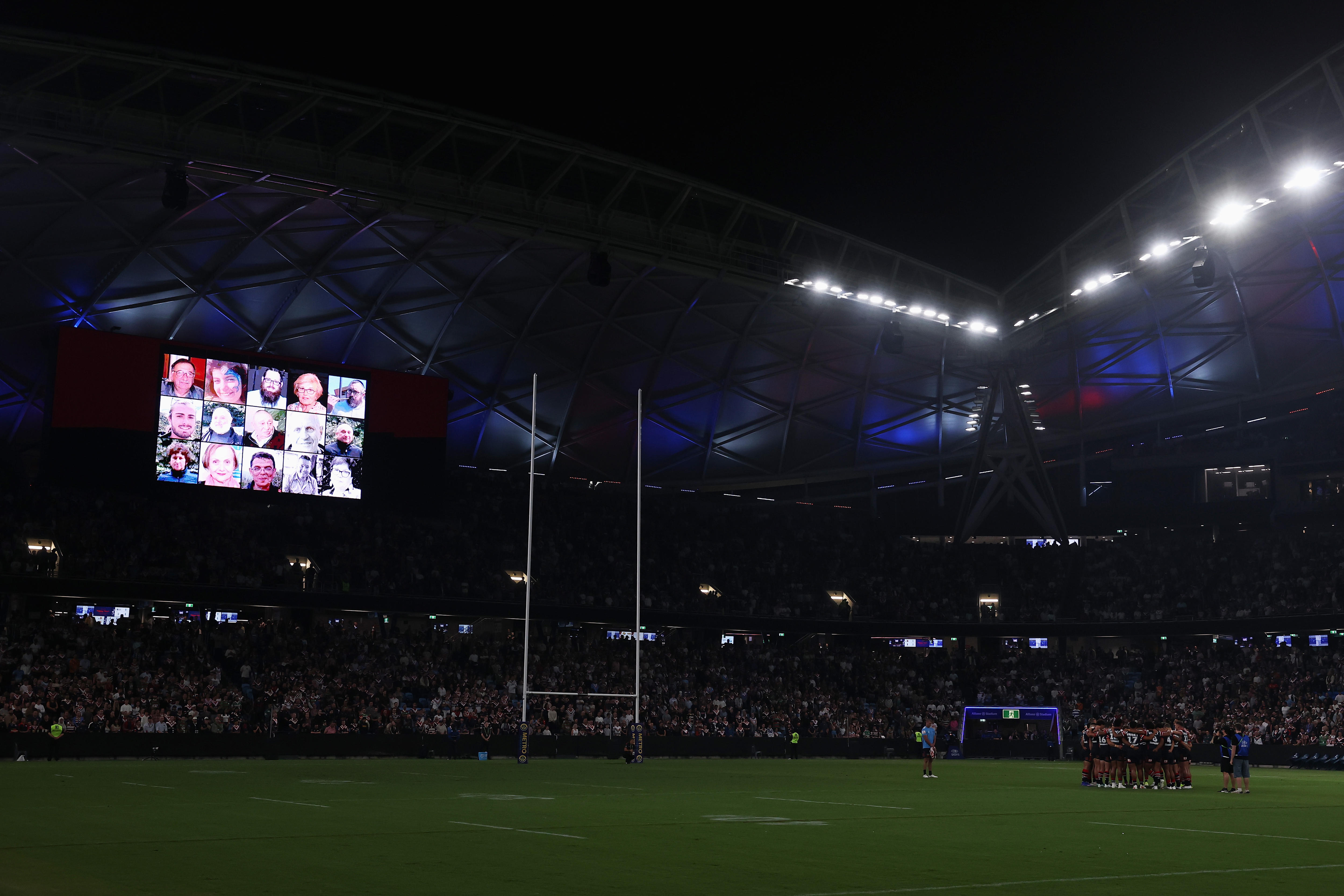Two teams of rugby league players stand on a dark field with photos of people on the big stadium screen