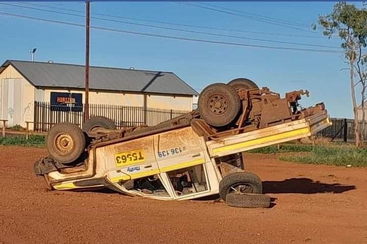 A white ute is upside down with a crushed roof on red dirt