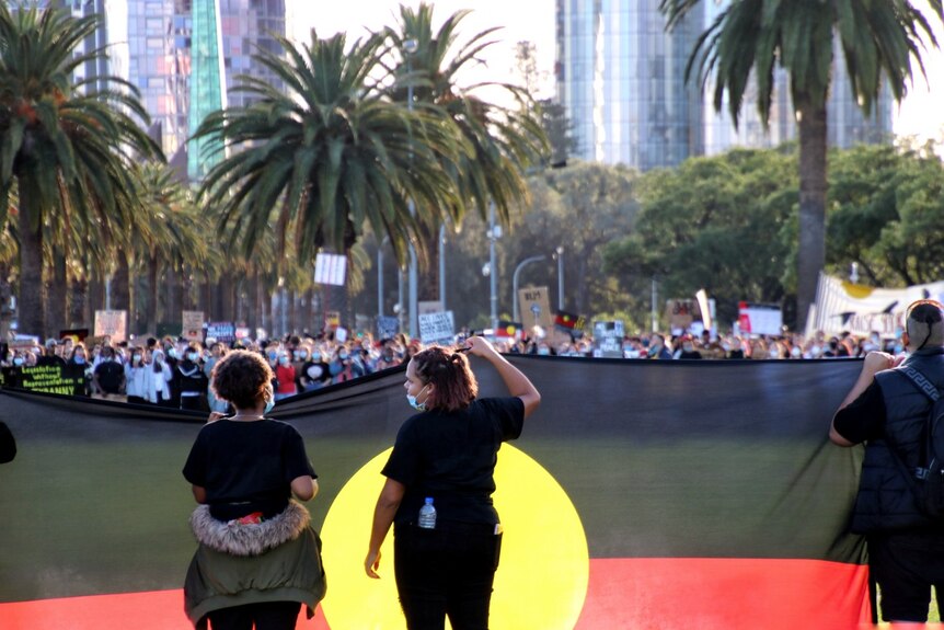 Two women stand side by side holding an Aboriginal flag in front of a crowd in the Perth CBD.