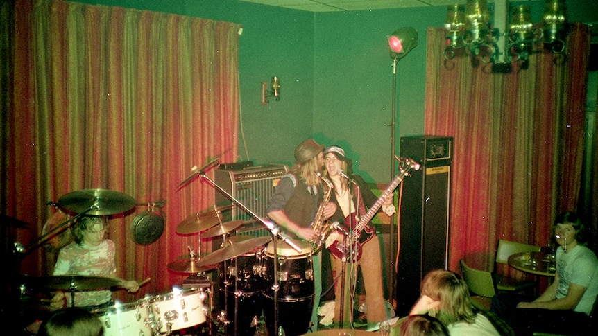 A band set up and playing in the corner of a crowded room, with red velvet curtains and green walls.