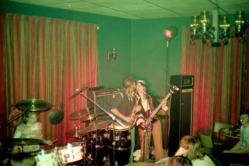 A band set up and playing in the corner of a crowded room, with red velvet curtains and green walls.