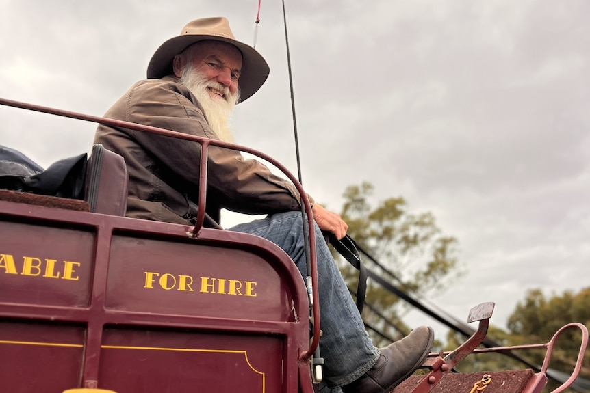 A man with a long white beard sits aboard a maroon stagecoach with a crop