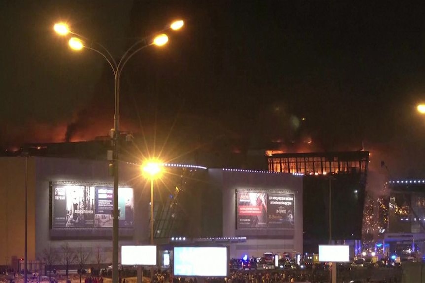 Concert hall in flames at night with hundreds of people gathered in the outside carpark and dozens of emergency vehicles. 