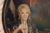 A woman in her late 60s with blonde hair and wearing a sparkly outfit, holds a glass of champagne and looks into a mirror