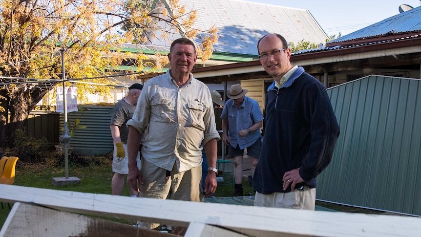 Two men standing next to a pile of wood with others working in the background