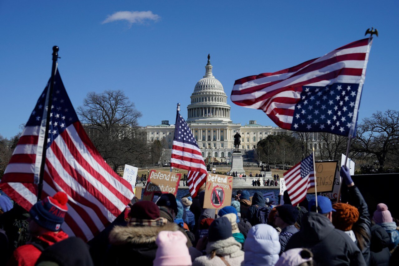 A large grand building is visible through a crowd of protesters, many of whom are holding signs and American flags