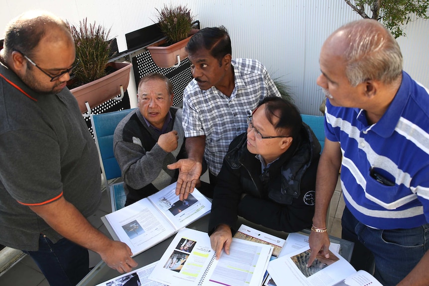 A group of grumpy men stand around a table with papers on it gesticulating at each other.