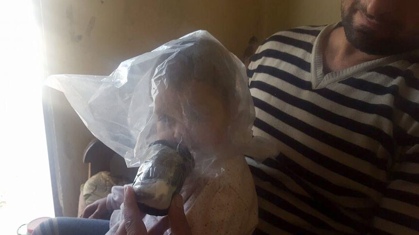 A baby sits on her father's lap wearing a plastic home made gas mask over her head.
