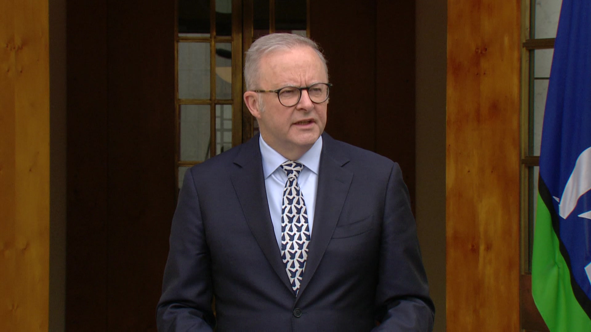 Albanese, vestido de terno, fala na porta do Parlamento. À direita você pode ver uma bandeira das Ilhas do Estreito de Torres.