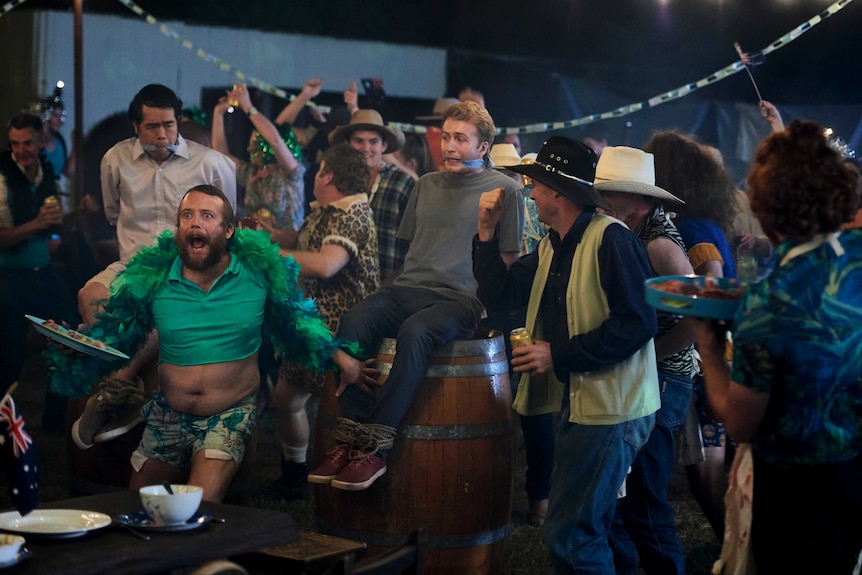 Around 20 people in this photo, mostly men wearing rural clothing and large hats. The main character sits gagged on a barrel.