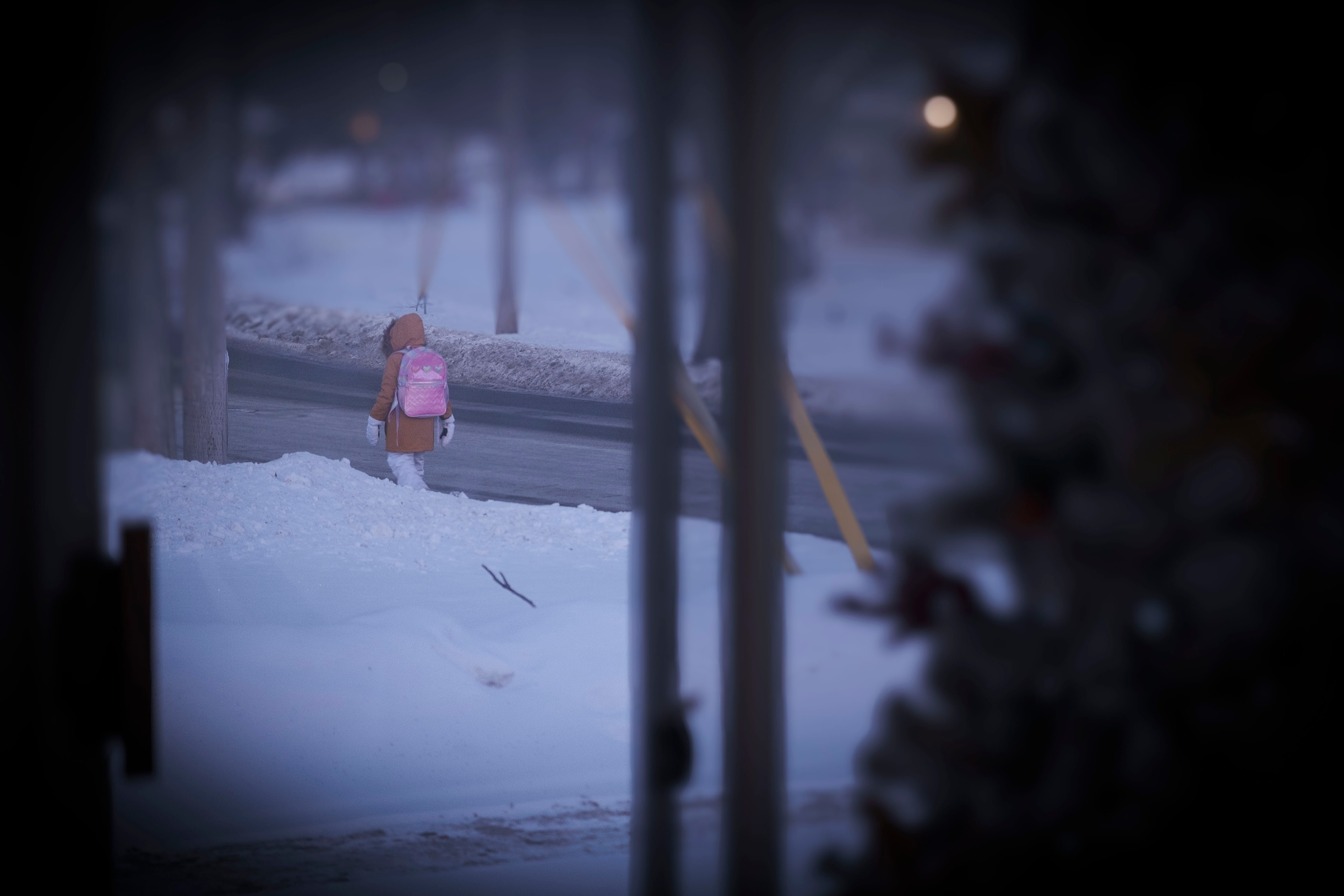 A child with a pink backpack walks down a street with snow beside the road.