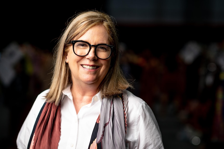 A woman looks at the camera and smiles. She wears dark glasses and has blonde hair.