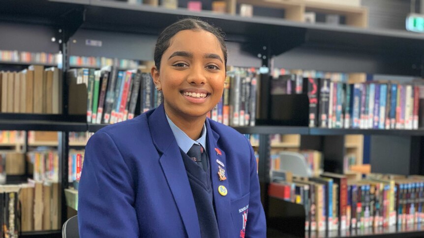 Nishadi sits smiling at the camera in front of a bookshelp, she wears a blue blazer and school uniform.