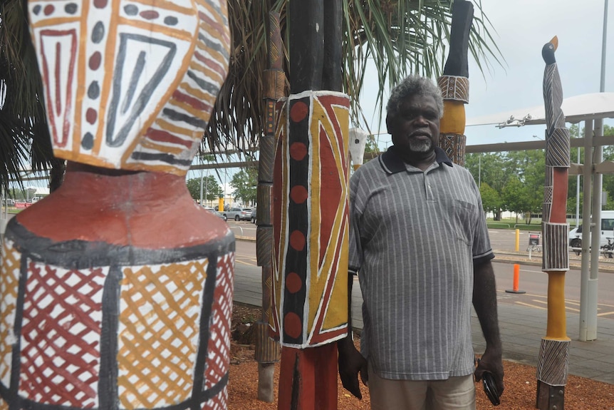 Yingiya Guyula stands next to an Aboriginal designed pole at Darwin Airport.