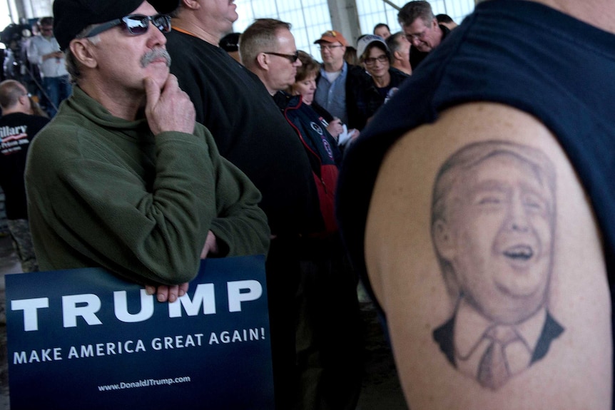Tattoo of Donald Trump's face on a supporter's upper arm with another supporter holding a sign reading TRUMP.