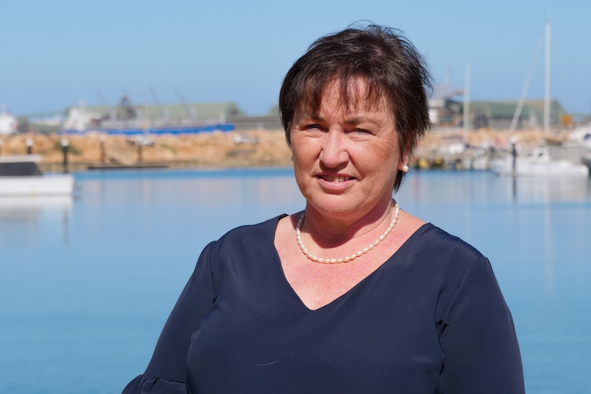 A middle-aged woman wearing a dark top and a pearl necklace stands in front of a harbour.