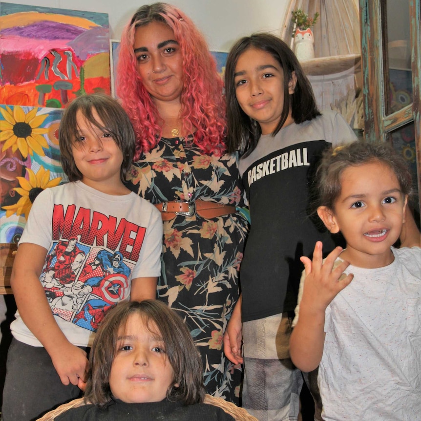 A woman with pink wavy hair stands behind four young boys