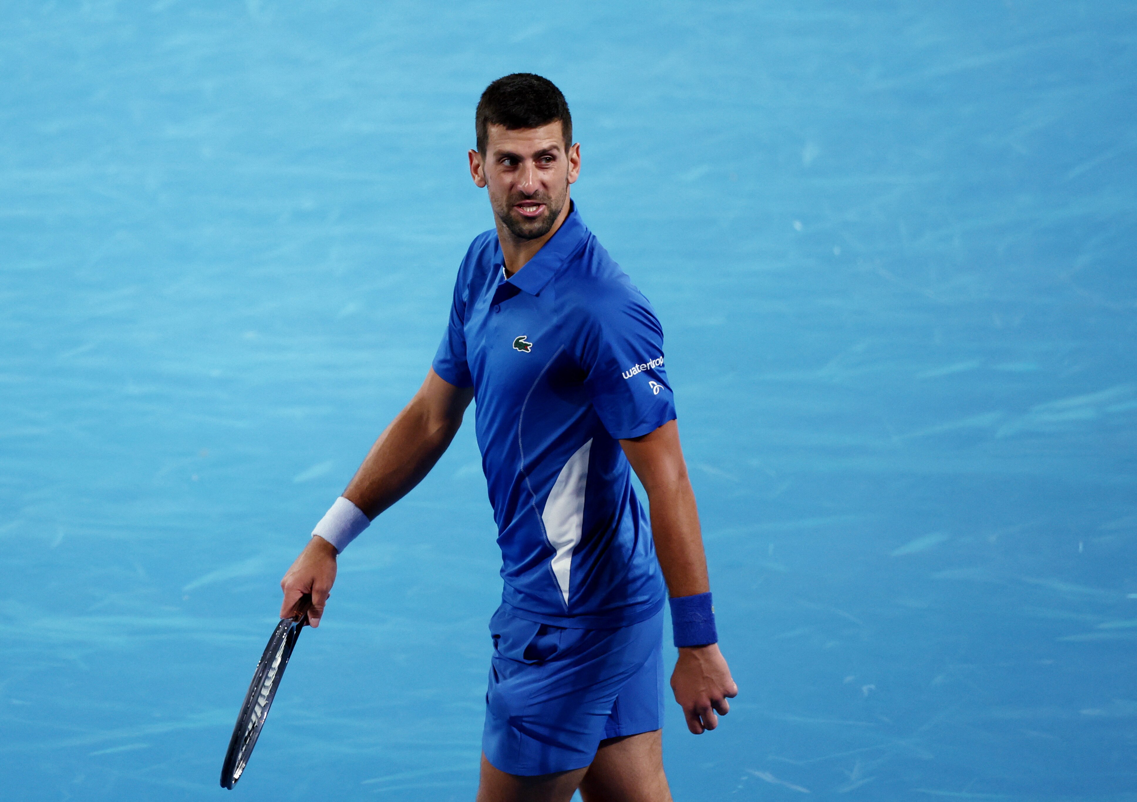 A male tennis player, holding his racquet, in all blue on a blue court, looks upset.