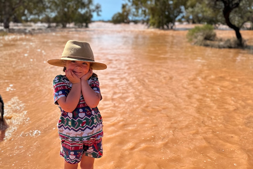 A little girl wearing a hat holding her face while standing in a muddy overflowing creek