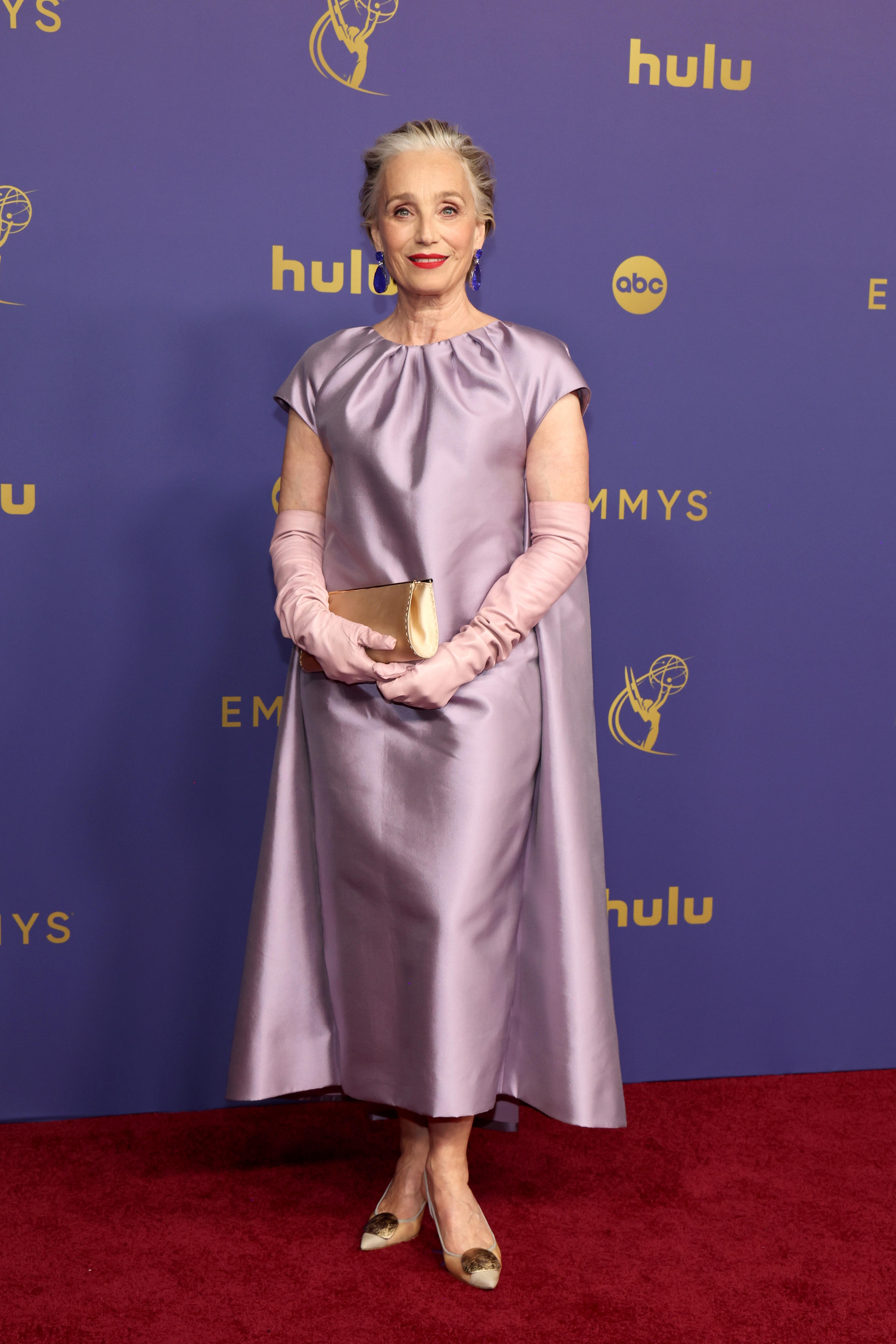 Dame Kristin Scott Thomas wearing a pale purple ankle-length dress with a cape, gloves and a gold clutch