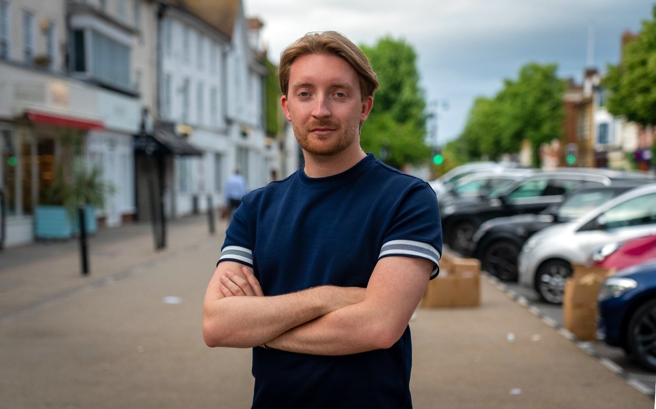 A man with his arms folded stands on the side of a road and smiles at the camera.