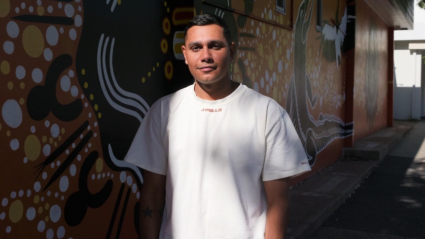 Rapper J-Milla stand in front of an orange mural in darwin, he wears a white shirt