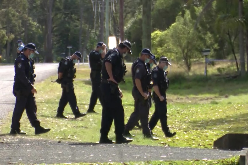 police in uniform in a row walk across a grass road verge with their heads down