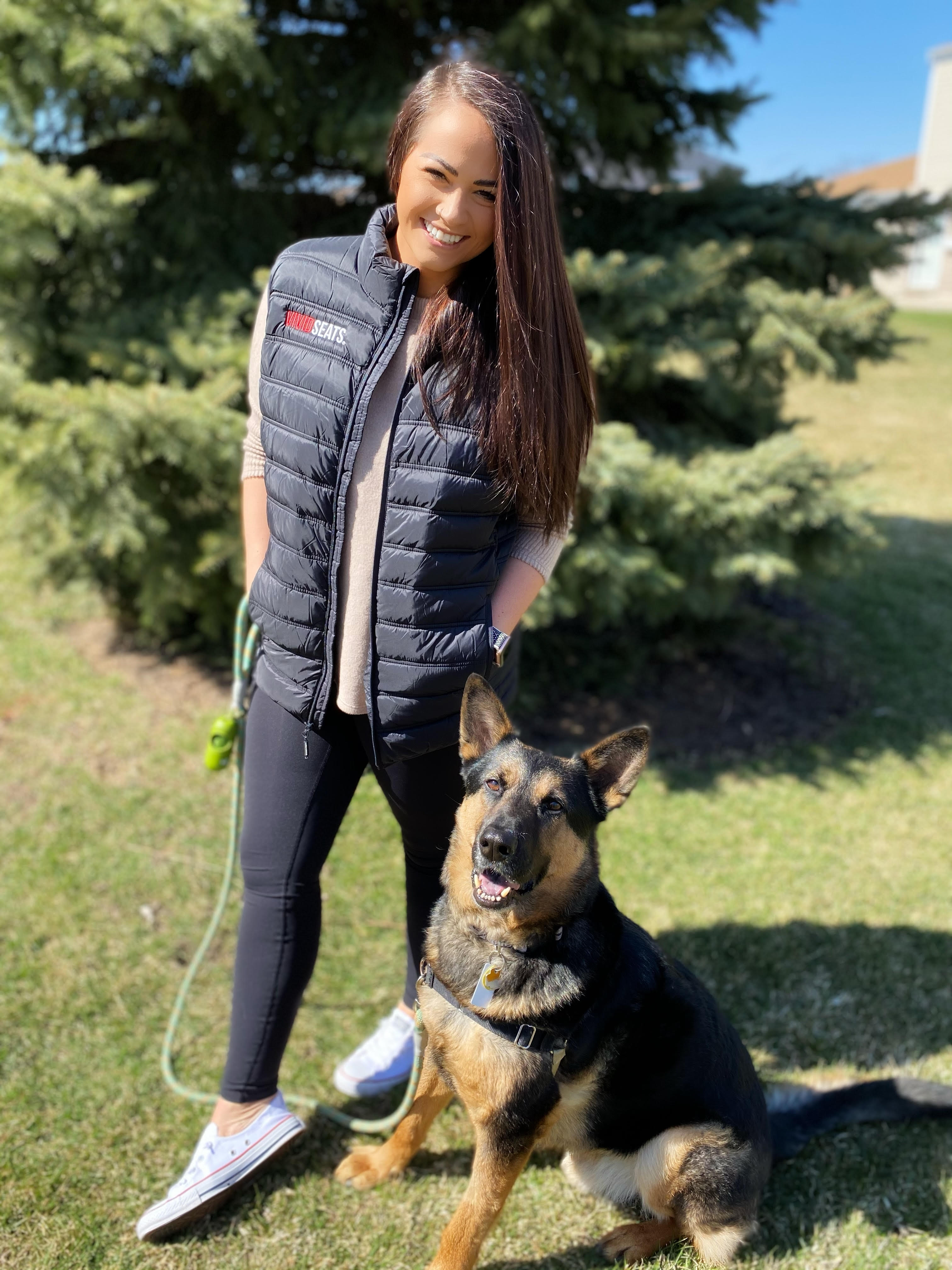 A young woman with long dark hair stands next to a German Shephard 