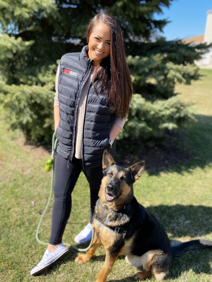 A young woman with long dark hair stands next to a German Shephard 