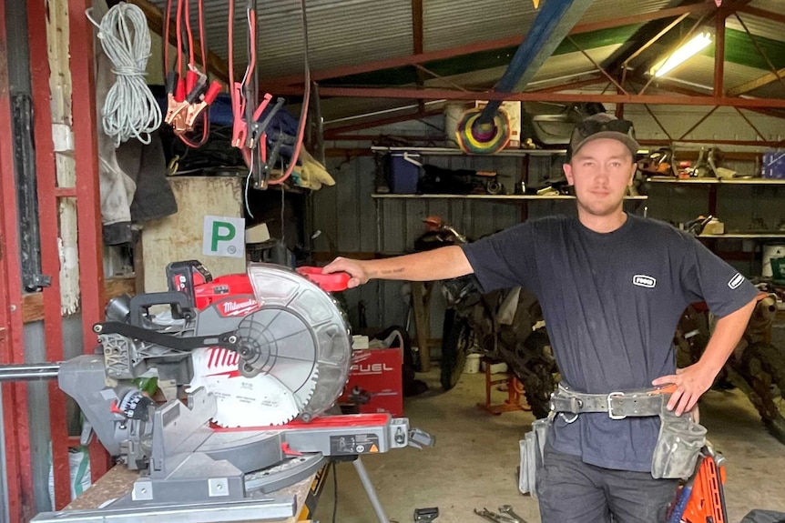 A young man stands in his workshop with power tools and motorbikes.