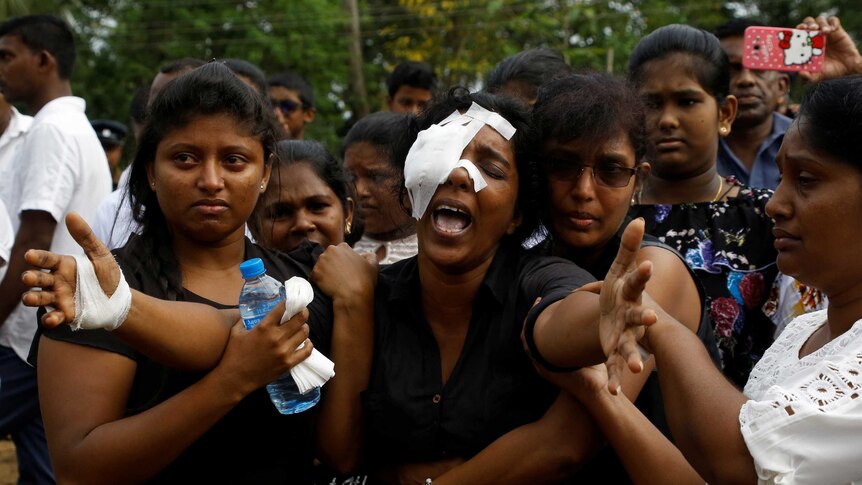 A woman with her right eye and hand bandaged spreads her arm wide and yells while people hold her arms and hug her