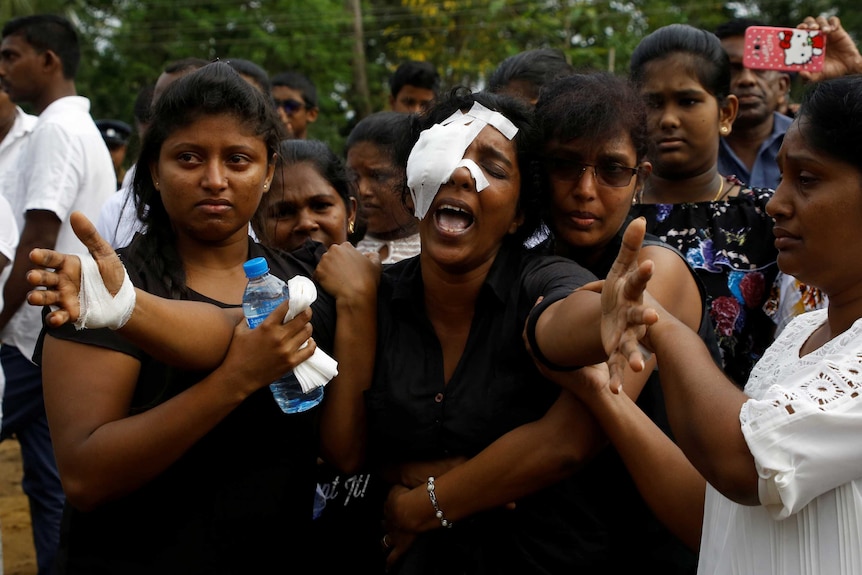 A woman with her right eye and hand bandaged spreads her arm wide and yells while people hold her arms and hug her