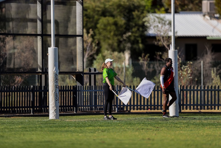An umpire, a woman, waves two white flags on the goal line of a football pitch.