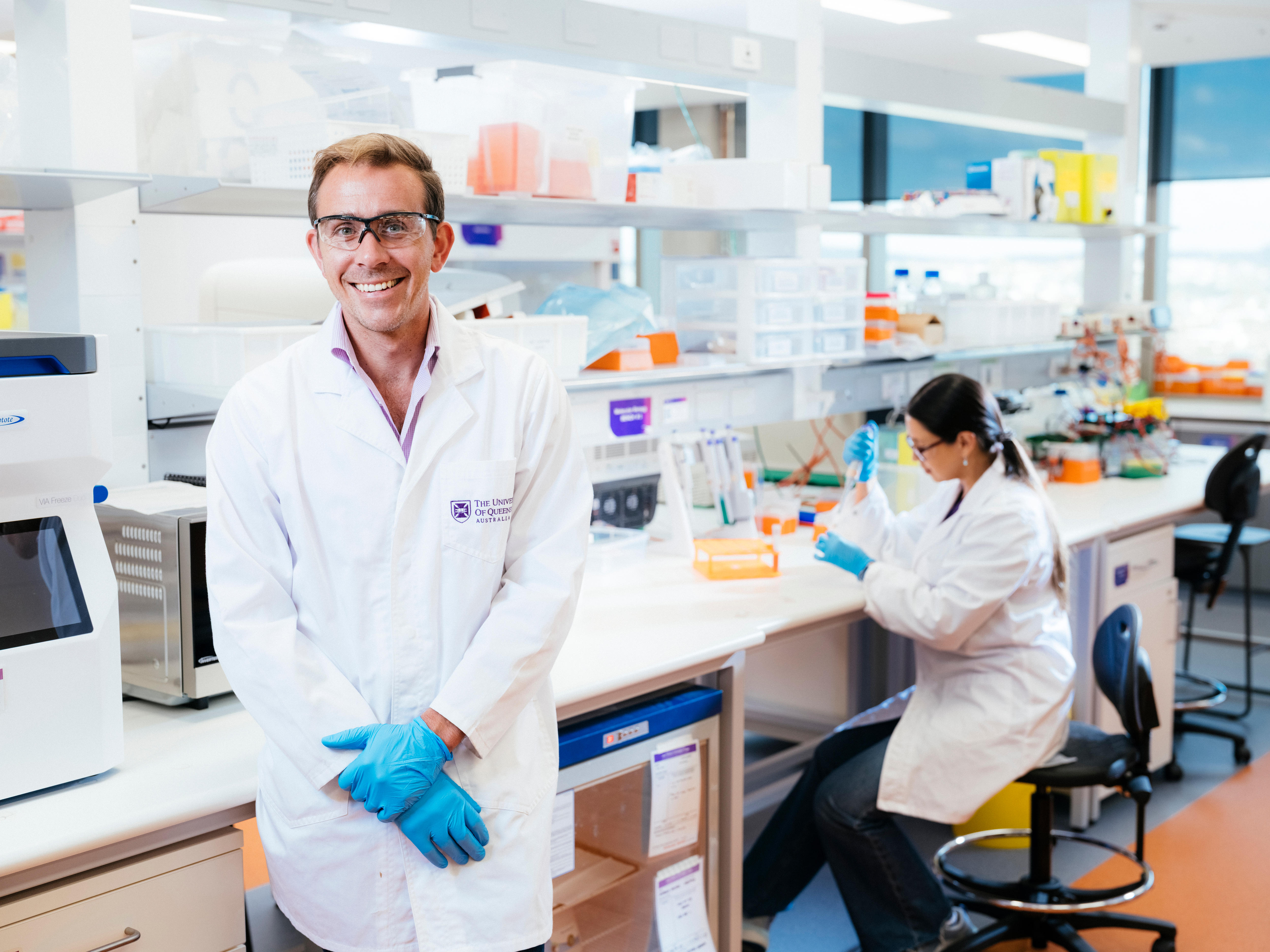 A man in a lab coat, gloves, and glasses smiling with a lab and lab tech in the background