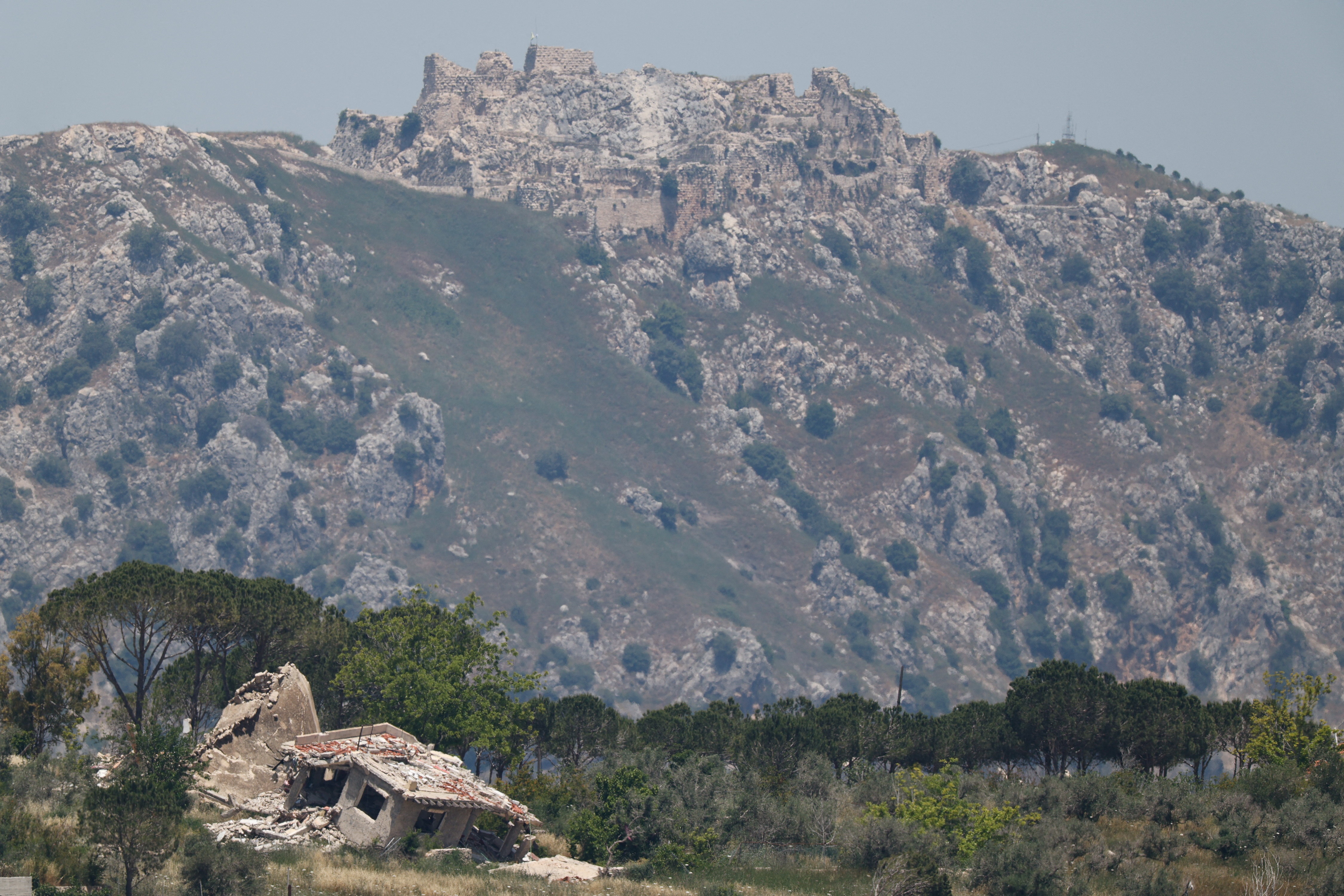 Ruinas derrumbadas de un castillo medieval en una enorme montaña salpicada de vegetación, detrás de un primer plano de árboles.