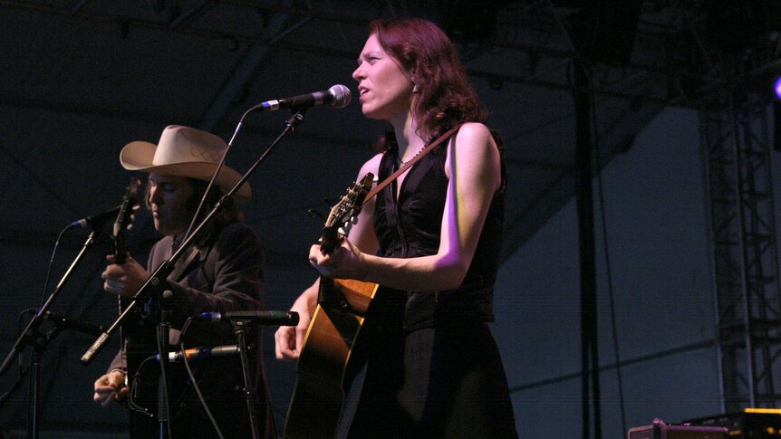 Gillian Welch and David Rawlings play guitars on stage. Rawlings wears a large cowboy hat.