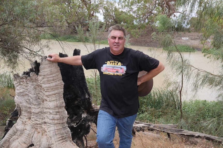 A man in a black shirt stands leaning on a hollowed out tree stump in front of a river.