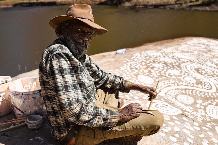 Keith Stevens sits upon a rock overlooking a waterhole, painting the surface with white paint.
