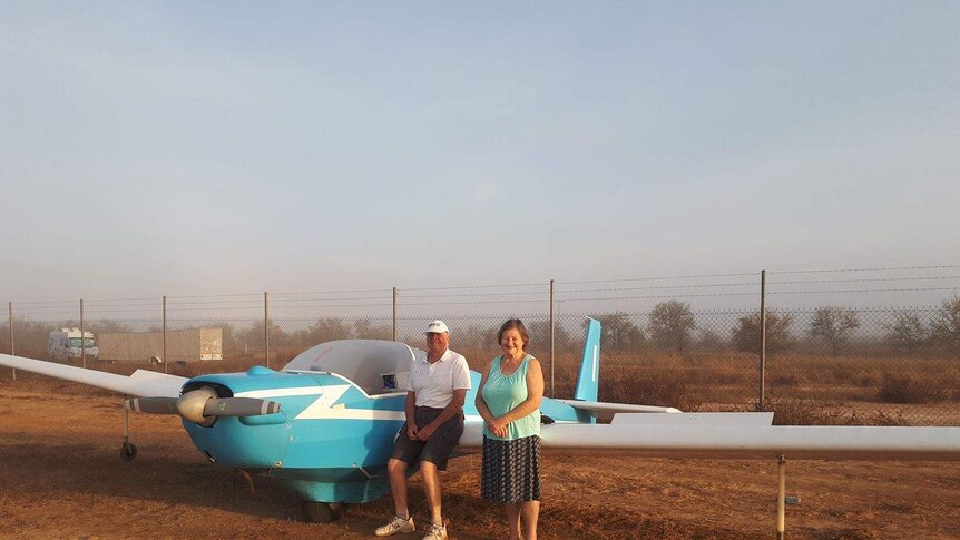 Glider pilot Graeme Clinton and friend Gloria leaning against a blue glider plane in the morning light.