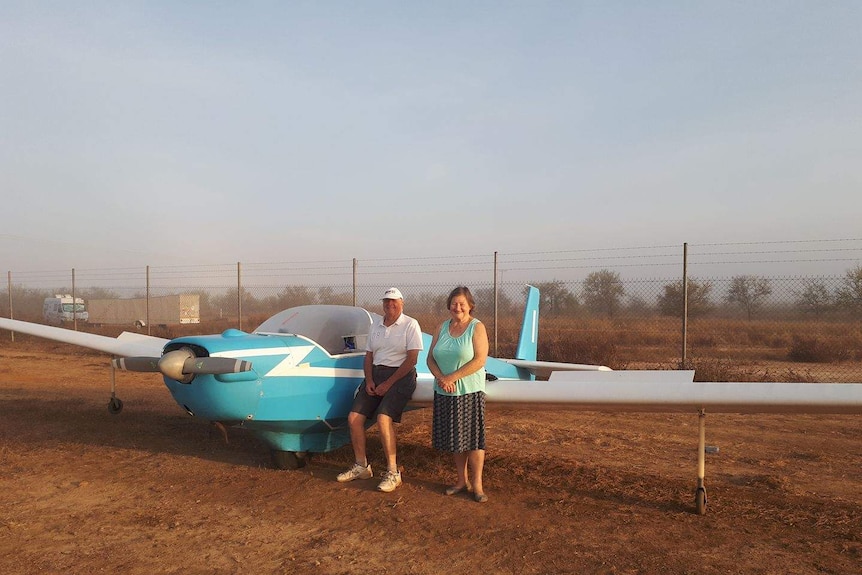 Glider pilot Graeme Clinton and friend Gloria leaning against a blue glider plane in the morning light.